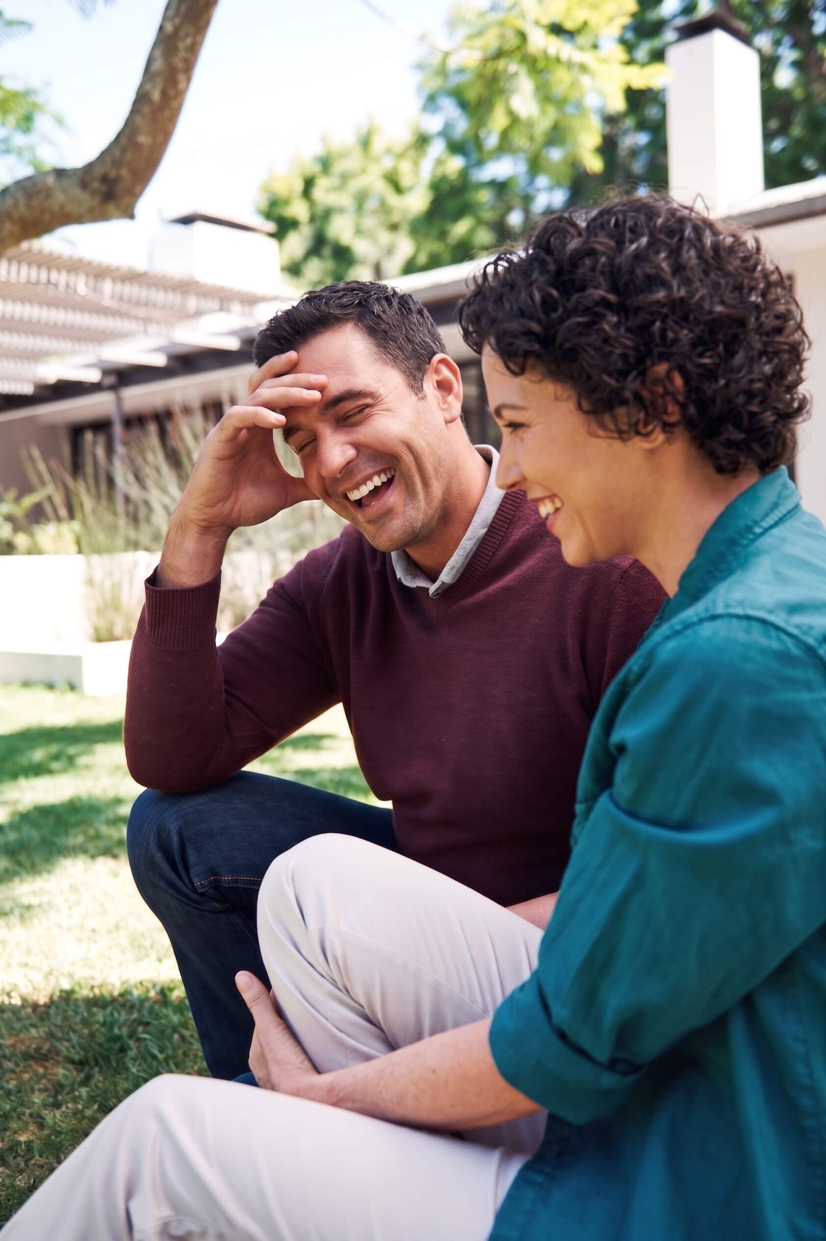 A couple sits outdoors on bright green grass, laughing together. The man, wearing a maroon sweater, rests a hand on his forehead as he smiles. The woman, in a teal shirt, smiles widely beside him. Their joyful interaction and relaxed posture reflect the vitality and calm of a regulated nervous system.